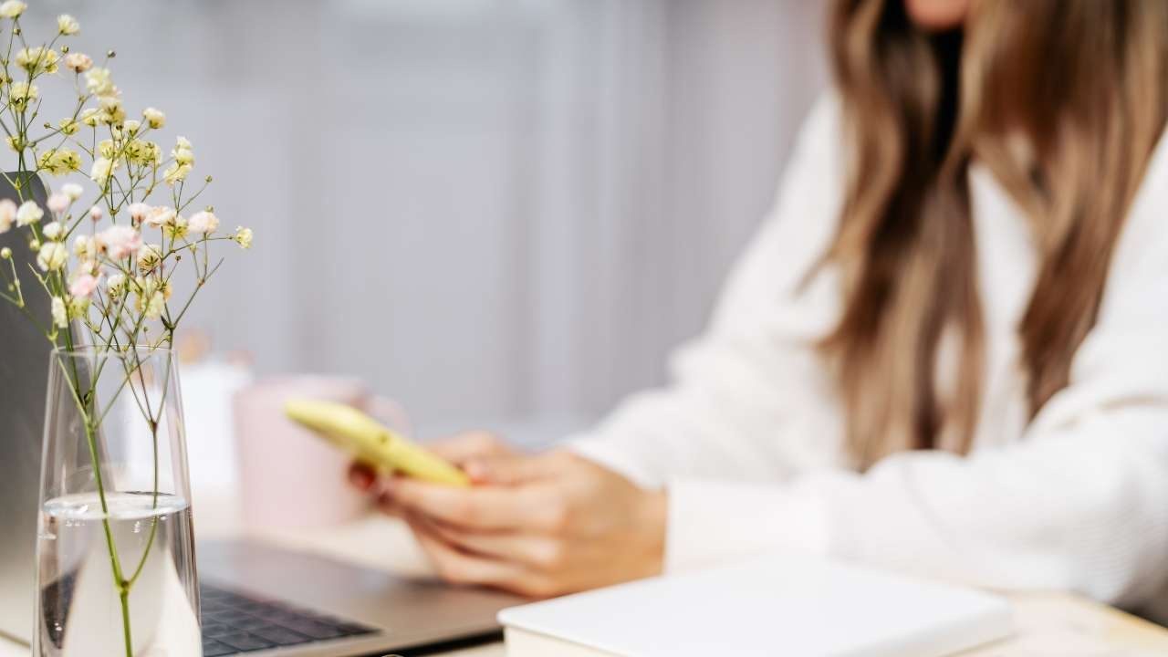 Женщина сидит за письменным столом и в телефоне ищет информацию, где взять выгодный кредит